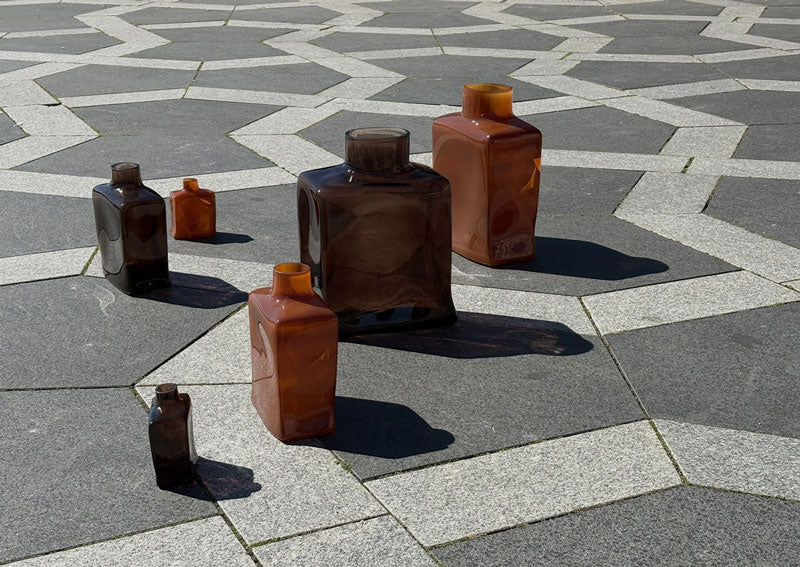 6 Shin glass vases in 3 sizes and 2 colours arranged on a gray and white geometric tiled pavement under bright sunlight.